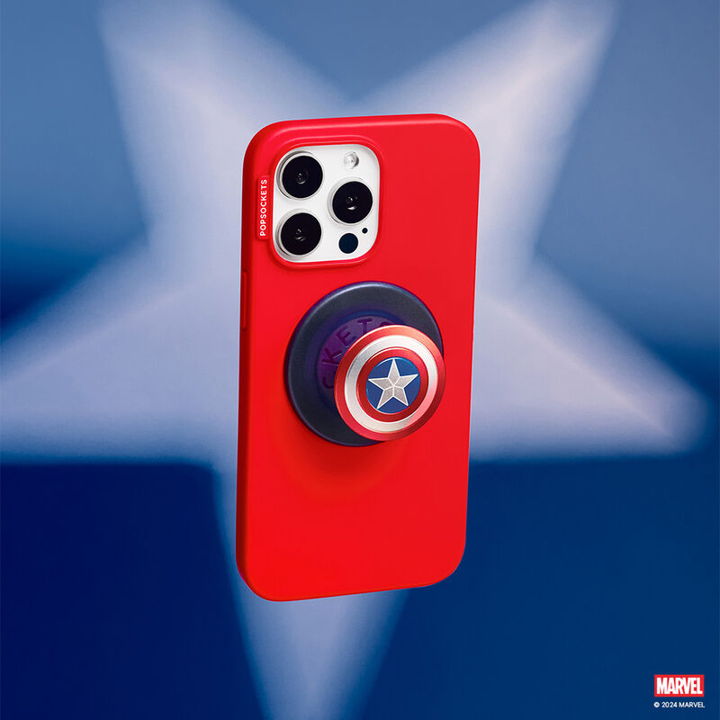 Red smartphone case with an Enamel Captain America PopGrip for MagSafe attached to the back. The PopGrip features Captain America's shield design with concentric red, white, and blue circles, and a white star in the center. The phone is shown from the back, positioned against a blurred background featuring a star shape.
