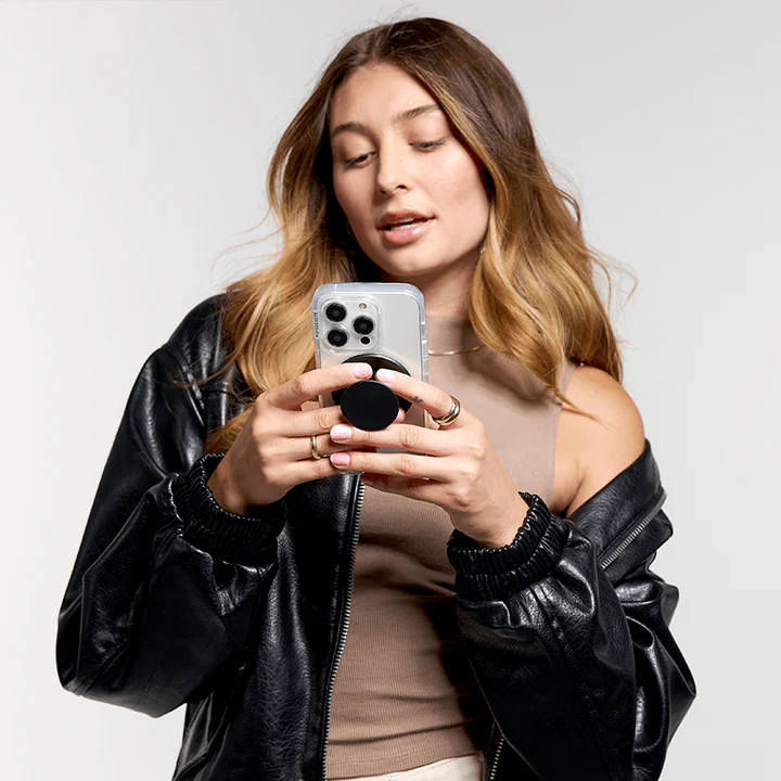 A woman with long, wavy hair is shown holding a smartphone with a clear case. The phone has a PopGrip MagSafe in black attached to its back. The woman is wearing a black leather jacket over a beige top and is looking at her phone, demonstrating the functionality and sleek design of the PopGrip while using her phone. The PopGrip is extended, highlighting its practical use as a grip for easier handling