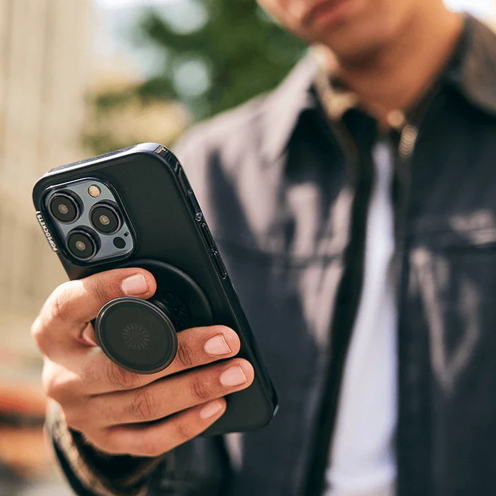 A man is shown outdoors, holding a smartphone with a black case. The phone has a PopGrip MagSafe in black attached to its back. The man is wearing a dark jacket and is looking at his phone, demonstrating the functionality and sleek design of the PopGrip while using his phone. The urban background suggests he is in a city setting. The PopGrip is extended, showcasing its practical use as a grip.