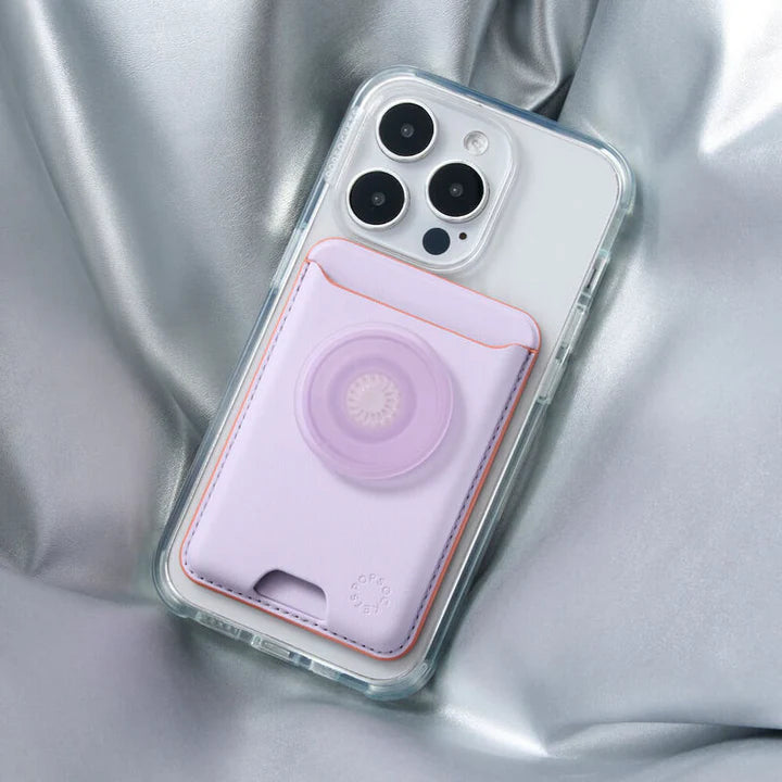 An iPhone with a clear case lying on a silver satin fabric. Attached to the back of the iPhone is a PopSockets MagSafe Softgoods PopWallet in Pale Lavender color. The wallet has a PopSocket in the same pale lavender shade, with an embossed logo near the bottom. The wallet's stitching is visible and provides a subtle contrast to the lavender color, enhancing the elegant and stylish look of the accessory.
