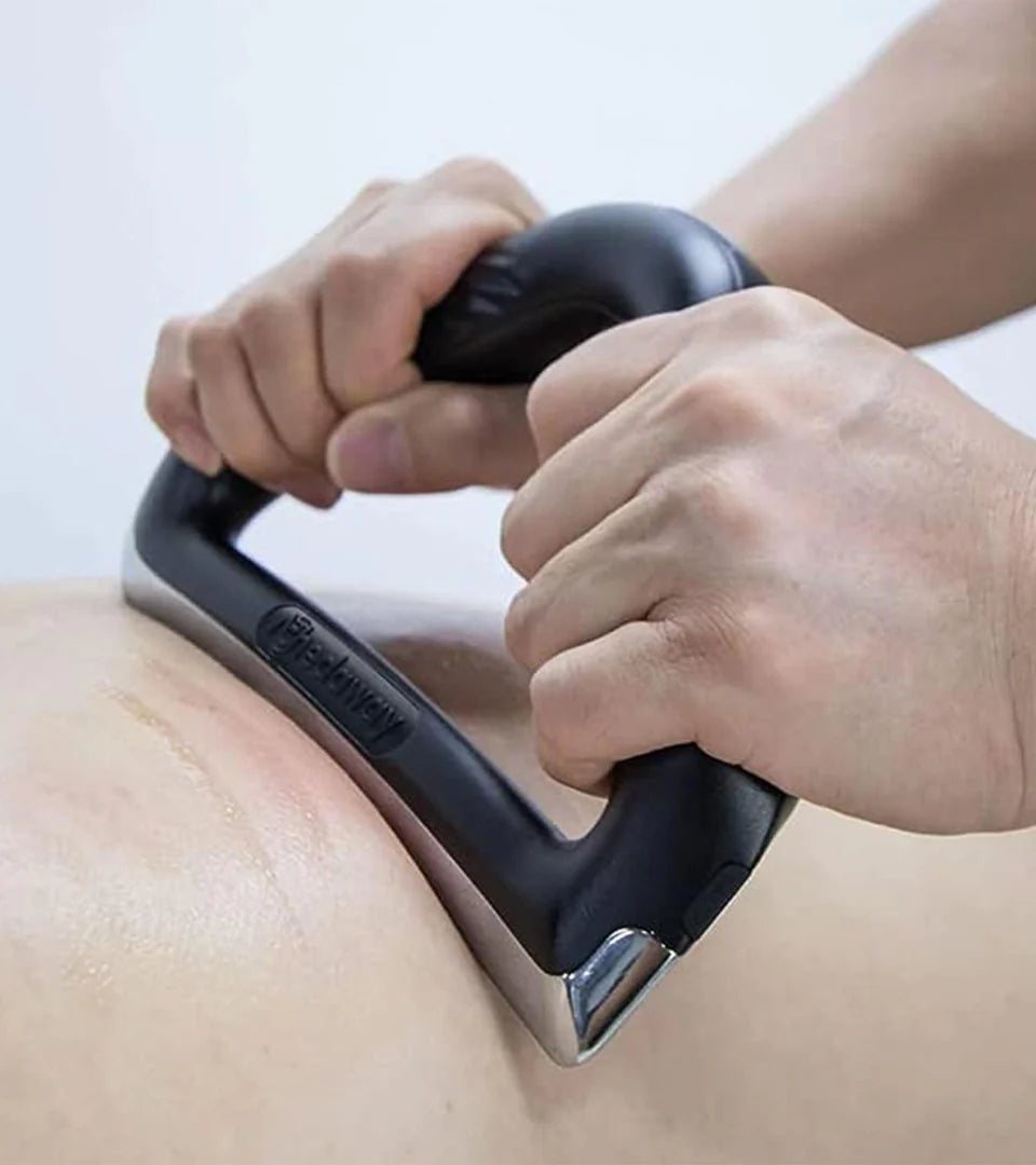 Person using a black and silver ergonomic massage tool on a bare back, applying firm pressure with both hands. The tool features a triangular design with a textured grip and an embossed brand name for enhanced control during muscle relief therapy.