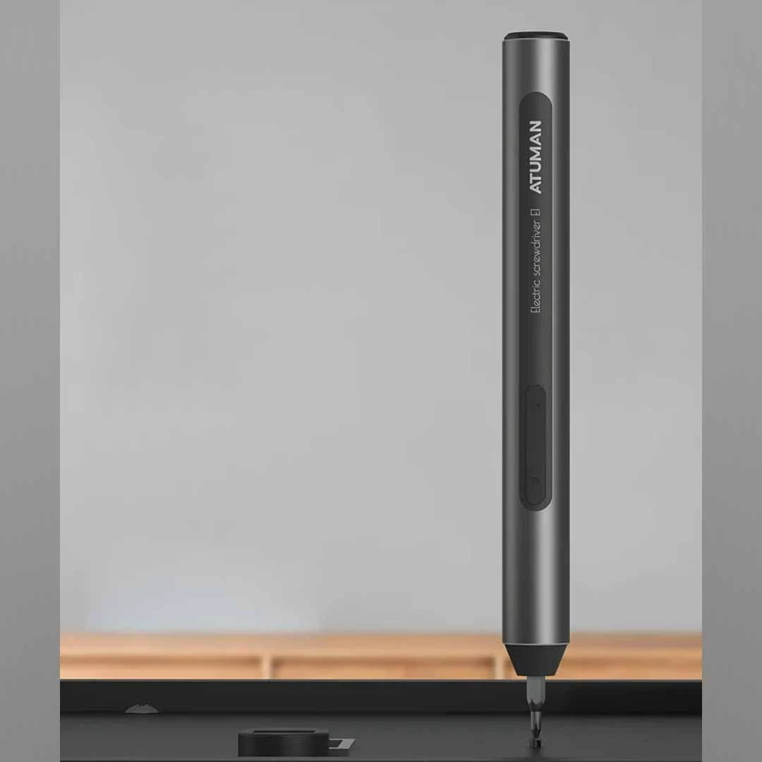 A black pen-shaped electric screwdriver branded with "ATuMan" is positioned vertically, with the tip pointed downward as if ready for use. The label reads "Electric Screwdriver ATuMan." The background is a blurred indoor setting, highlighting the tool's sleek and modern design.