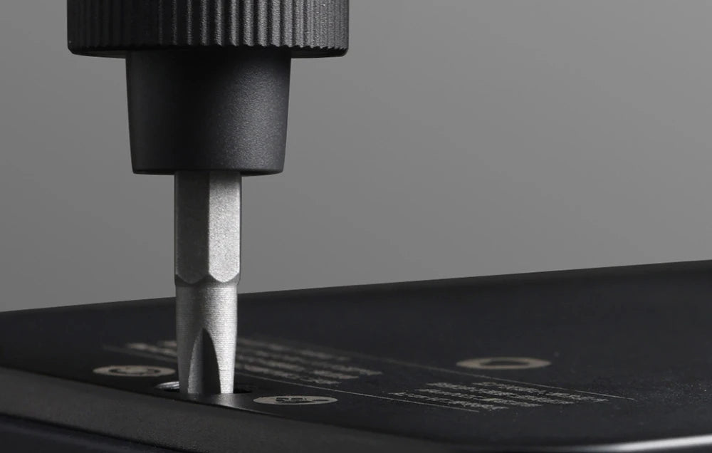 A close-up view of a screwdriver bit being used to tighten or loosen a screw on an electronic device. The handle of the screwdriver is black and textured for grip. The bit is precisely fitted into the screw head, demonstrating the tool's functionality. The background is a neutral gray, emphasizing the focus on the tool and the screw.