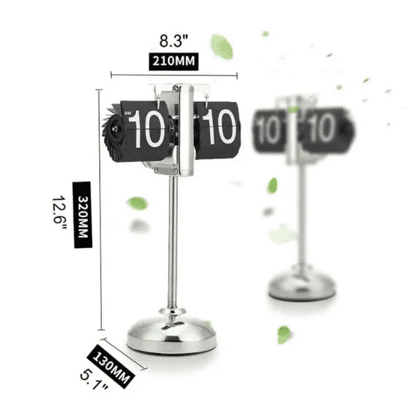 A vintage-style flip clock with a chrome stand is shown with its dimensions labeled. The height is 12.6 inches (320mm), the width is 8.3 inches (210mm), and the base diameter is 5.1 inches (130mm). The clock displays the time "10:10" with black and white flip cards. Another identical clock is blurred in the background, and a few green leaves are scattered around the image, adding a touch of natural decoration.