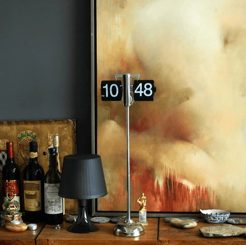 A vintage-style flip clock with a chrome stand displays the time "10:48" and is placed on a wooden table. The table also features various decorative items, including bottles of wine, a small black table lamp, a statue, and a small dish. In the background, there is a large abstract painting with warm tones of beige, brown, and red, adding a stylish and artistic ambiance to the setting.