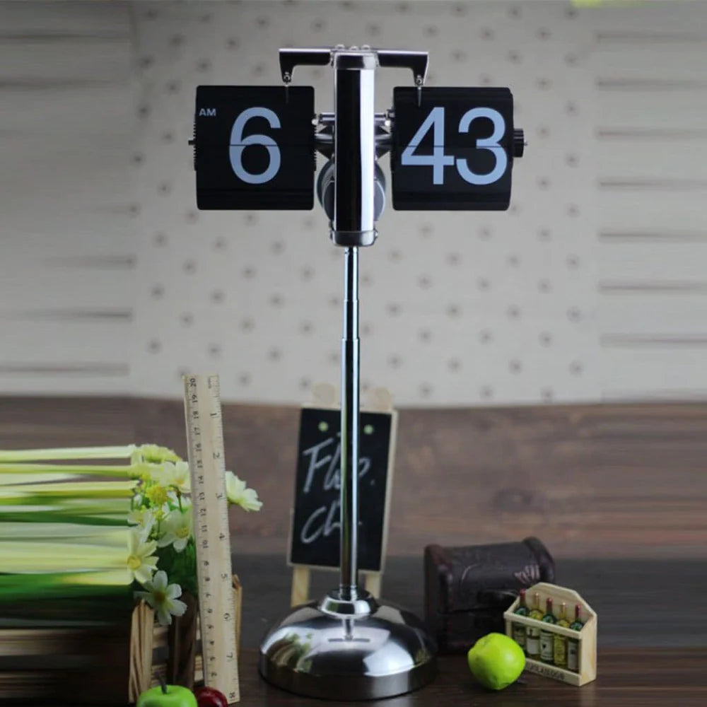 A vintage-style flip clock with a chrome stand displays the time "6:43 AM" and is placed on a wooden table. The table also has decorative items, including a bundle of artificial white flowers, a wooden ruler, a small chalkboard sign, a miniature crate with tiny bottles, a green apple, and a small rolled-up scroll. The background features a light-colored wall with subtle patterns, creating a charming and rustic ambiance.