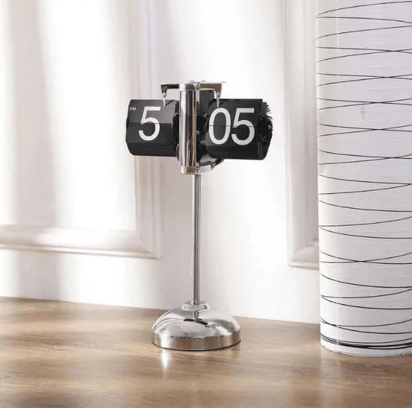 A vintage-style flip clock with a chrome stand is placed on a wooden surface. The clock displays the time "5:05" with its black and white flip cards. Next to the clock, there is a cylindrical decorative item with a white and black wavy pattern. The background features white walls and window trim, creating a clean and minimalist setting.