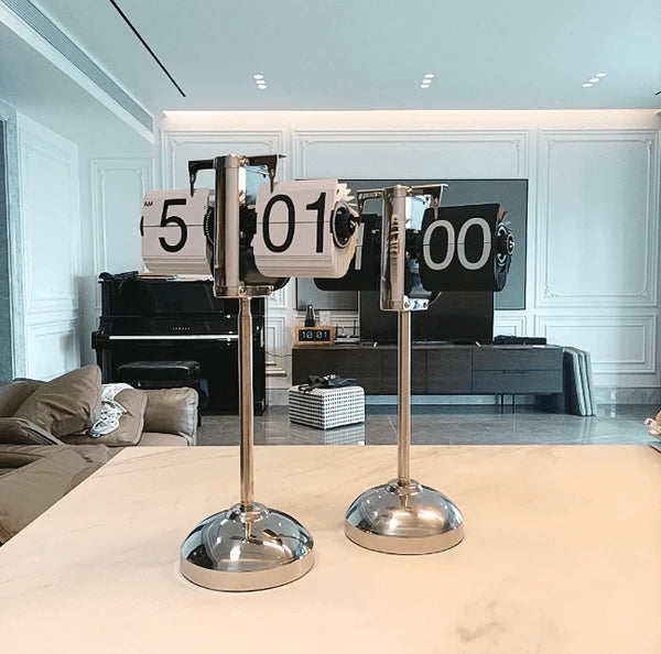 Two vintage-style flip clocks with chrome stands are placed on a light-colored surface. The clock on the left displays "5:01," and the clock on the right displays "10:00." The background features a modern, well-lit living room with white walls, recessed lighting, a piano, and a large entertainment center. The setting is elegant and contemporary, with a mix of classic and modern elements.