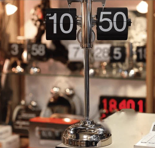 A vintage-style flip clock with a chrome stand displays the time "10:50 AM." The clock is prominently featured in the foreground. The background is a cluttered display of various other clocks and timepieces, both digital and analog, creating a busy and eclectic setting. The clocks are arranged on shelves and tables, showcasing a variety of designs and styles.