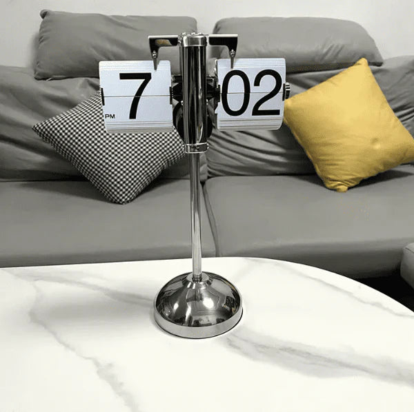 A vintage-style flip clock with a chrome stand is placed on a white marble surface. The clock displays the time "7:02 PM" with its white and black flip cards. In the background, there is a gray sofa with two cushions: one yellow and one black and white checkered. The setting is modern and stylish, with a clean and minimalist aesthetic.