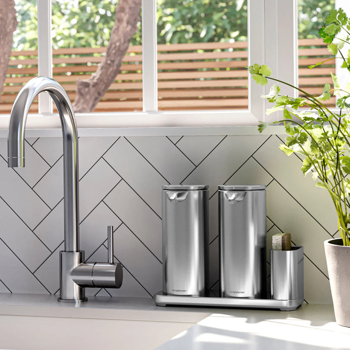 A bright and modern kitchen scene with a stainless steel faucet featuring a high, curved spout. Next to the faucet, there is a stainless steel kitchen organizer set that includes two tall, cylindrical containers and a compartment for a sponge. The backsplash is designed with white tiles arranged in a herringbone pattern. A potted plant with green leaves adds a touch of nature to the space, which is illuminated by natural light streaming in from a window that overlooks an outdoor area with trees and a woode
