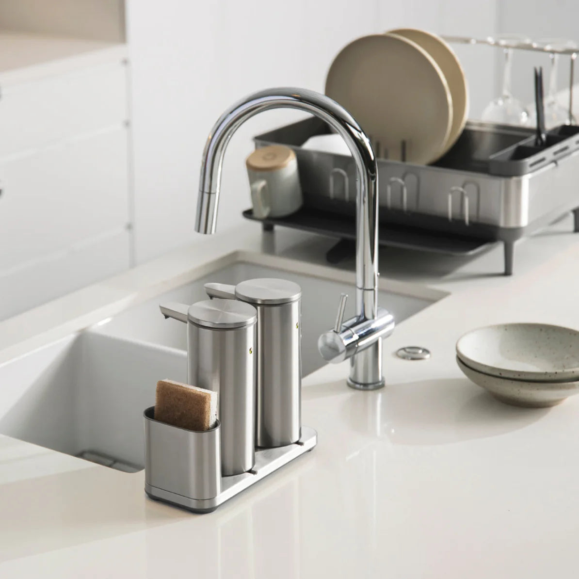 A modern kitchen sink setup featuring a sleek, stainless steel faucet with a high, curved spout. On the countertop beside the sink, there is a stainless steel organizer holding two cylindrical containers, likely for soap or lotion, and a sponge holder. The kitchen in the background has a minimalistic design, with a dish rack holding clean dishes, including plates and glassware, and a few bowls on the countertop, all contributing to an organized and contemporary kitchen environment.