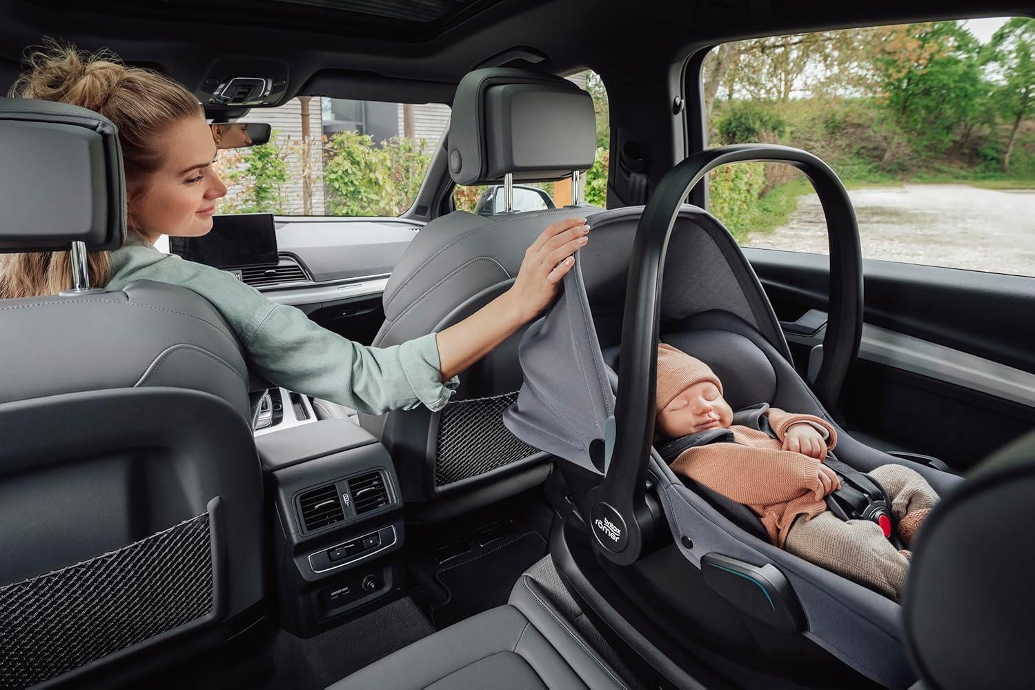 Mother sitting in the backseat of a car gently adjusting the canopy of a Britax Römer infant carrier, where a sleeping baby is securely fastened, highlighting comfort and safety during travel.