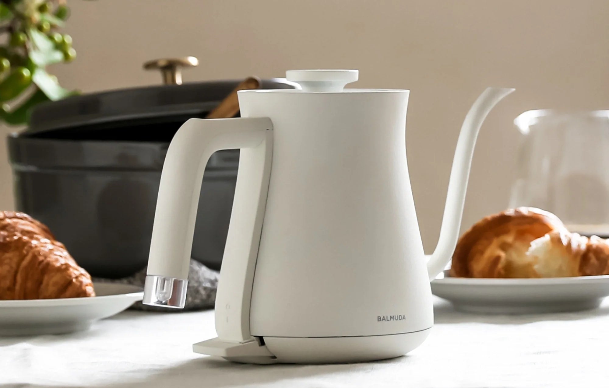 The image shows a Balmuda electric kettle in a sleek, matte white finish, placed on a light-colored countertop. The kettle features a minimalist design with a curved spout for precise pouring and an ergonomic handle with a clear, modern design element at the base.
