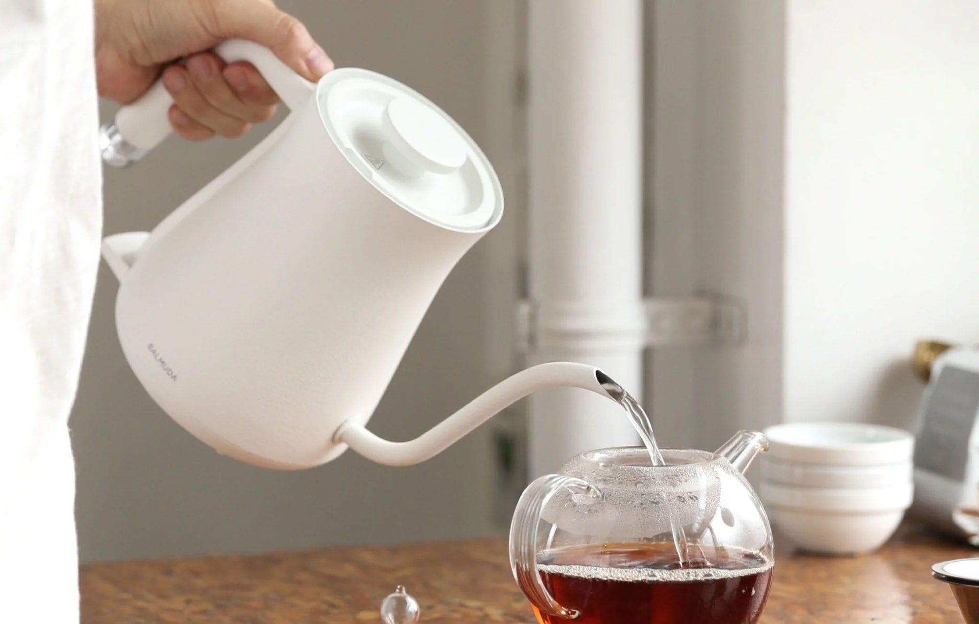 The image shows a person pouring hot water from a white, minimalist electric kettle into a clear glass teapot filled with tea. The kettle has a sleek, modern design with a long, narrow spout for precise pouring.