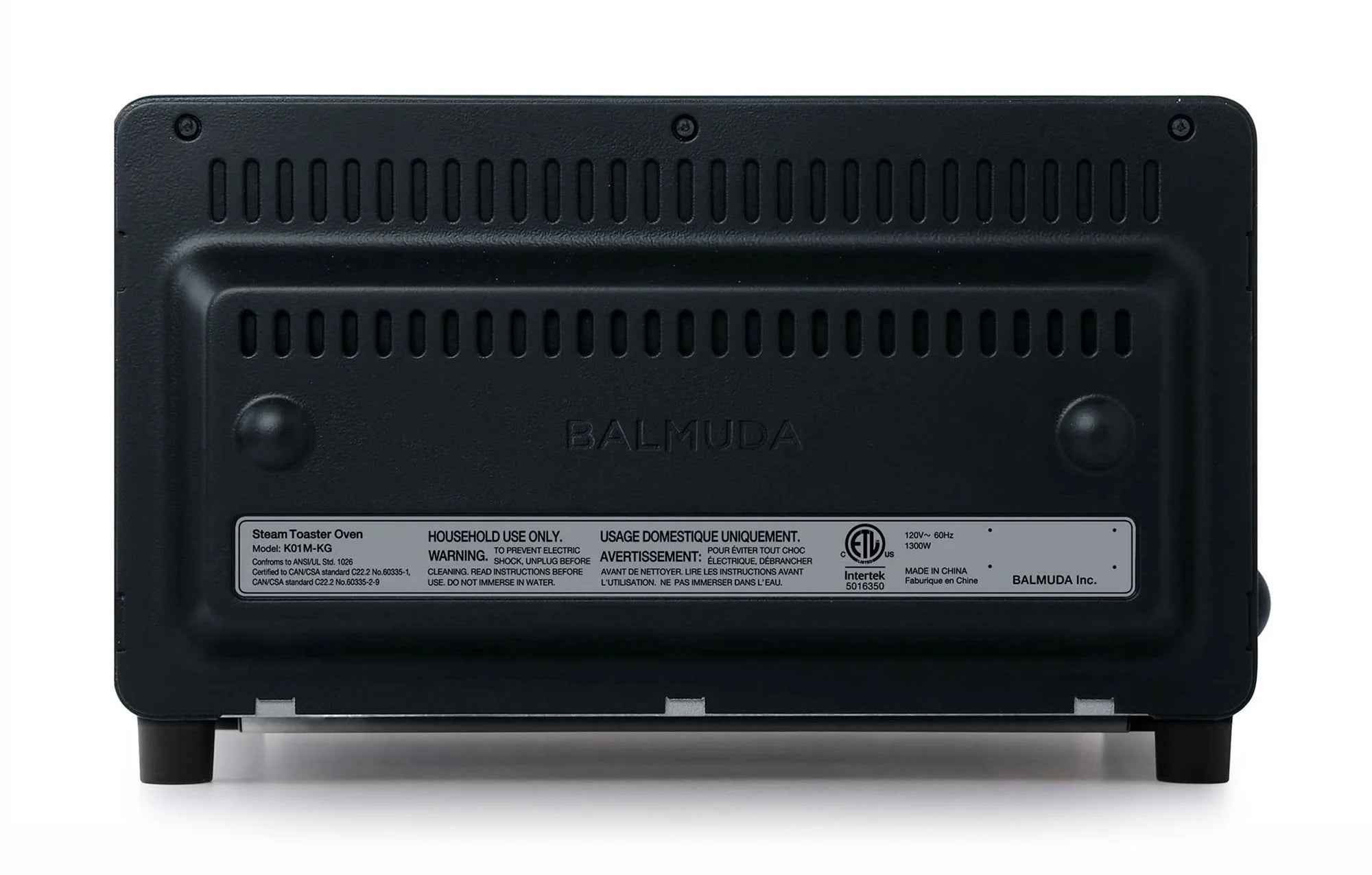 The image shows the back view of a Balmuda toaster in a matte black finish. The back panel features a series of horizontal ventilation slots near the top for airflow and cooling. The center of the panel has the brand name "BALMUDA" embossed into the surface. Below the brand name, there is a label displaying important safety and usage information, including warnings and certification details.