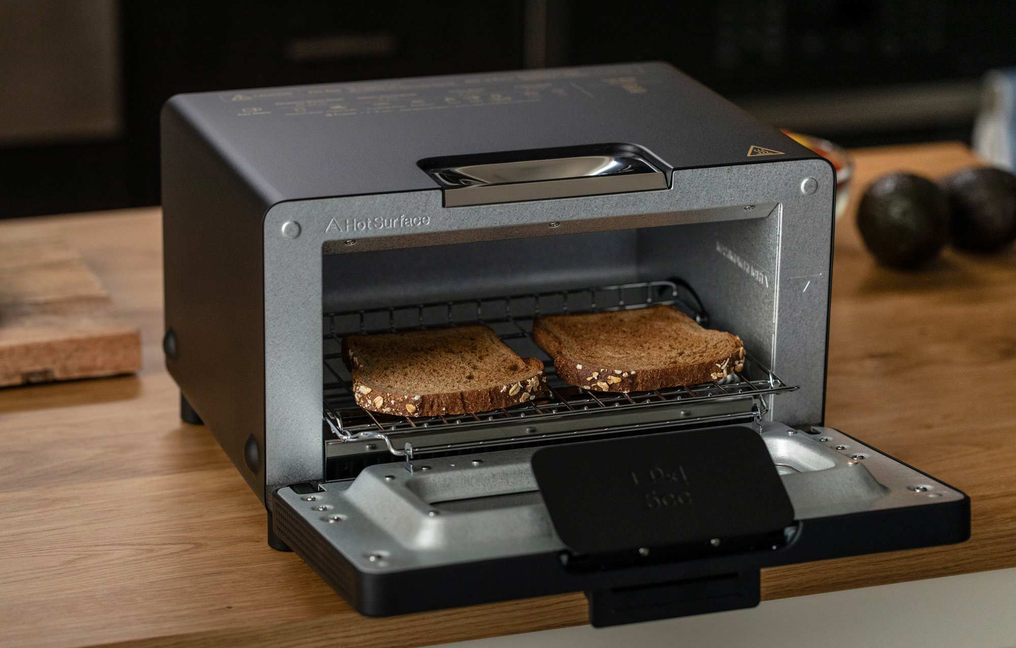 The image shows a Balmuda toaster in a matte black finish with its door open, revealing two slices of bread toasting inside. The toaster is placed on a wooden kitchen countertop, with a few kitchen items visible in the background, including a cutting board and some avocados. The interior of the toaster is lit up, highlighting the wire rack holding the bread slices