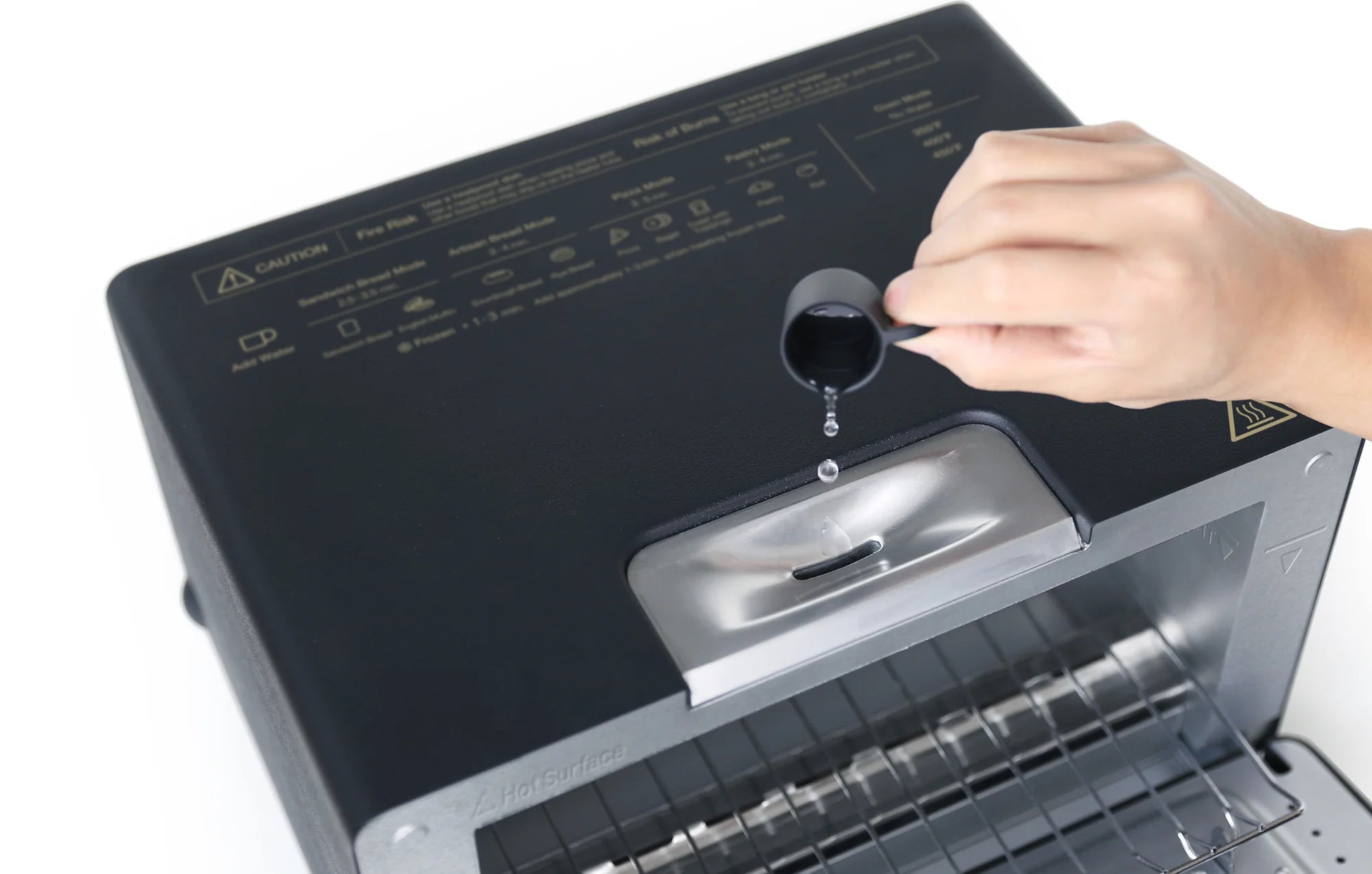 The image shows a close-up of the top of a Balmuda toaster, where a hand is seen pouring water into a small reservoir on the appliance's top surface using a measuring spoon. The toaster’s interior, including the wire rack, is visible through the open door.