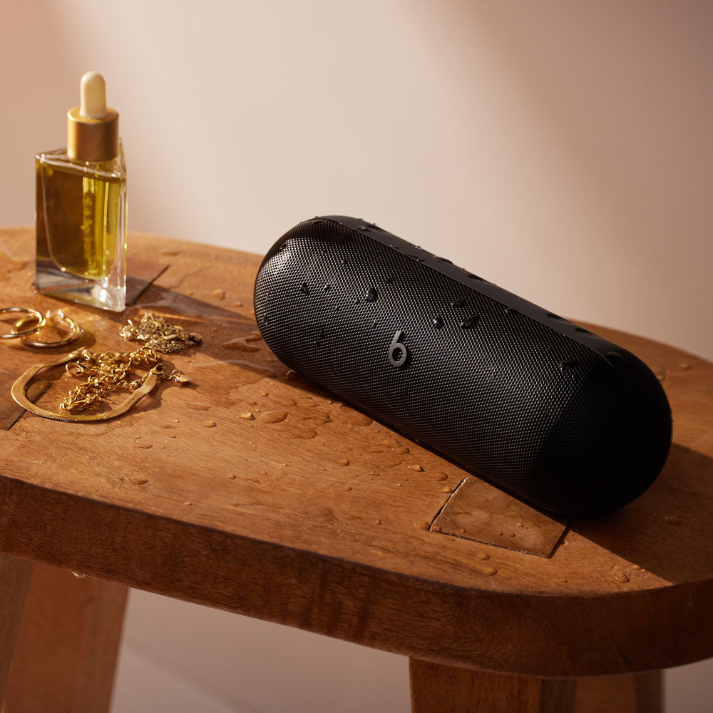 Alt text for this image:
A black Beats portable speaker with water droplets on its surface, sitting on a wooden table next to jewelry and a bottle of oil, highlighting its water-resistant feature and modern design in an elegant setting.