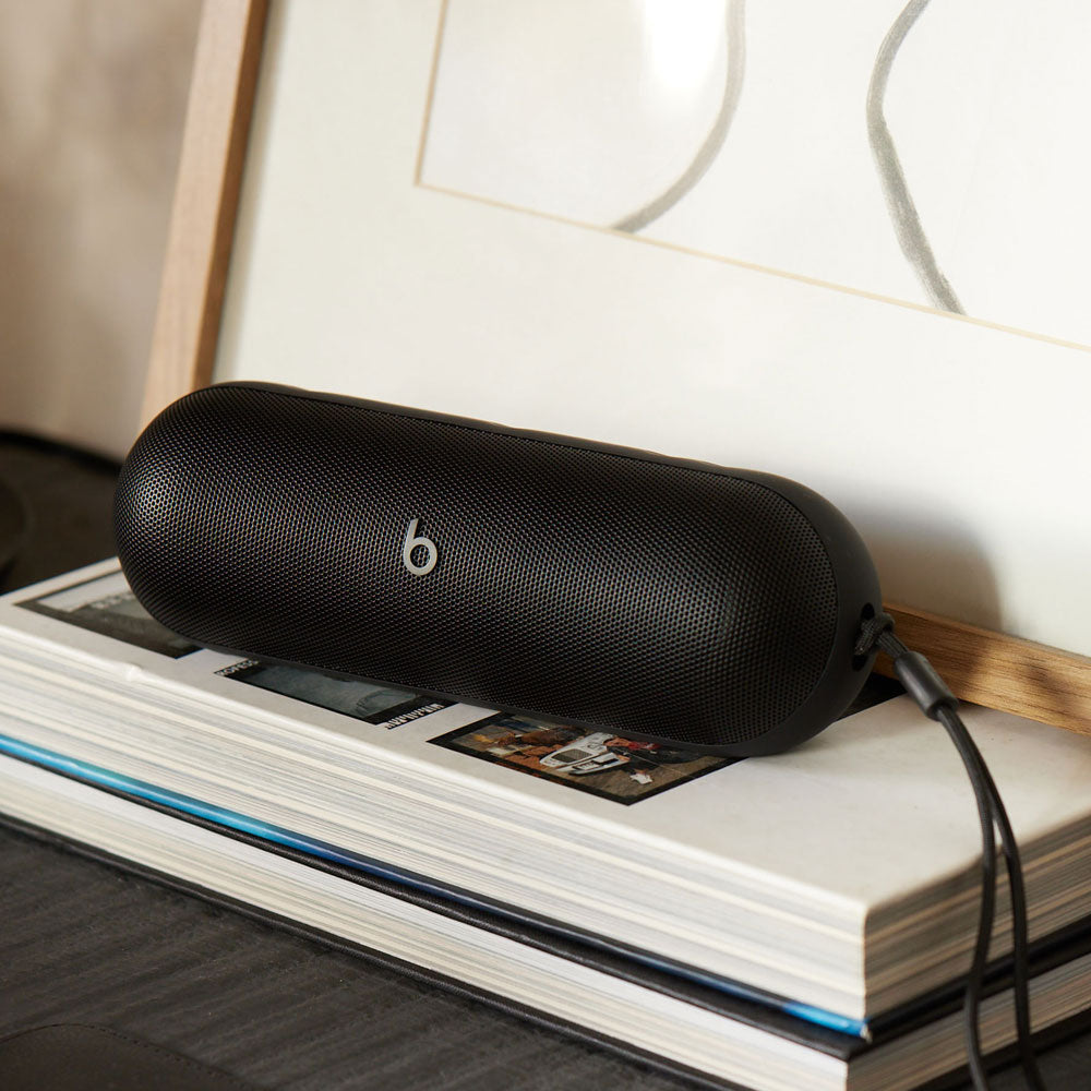 Alt text for this image:
A black Beats portable speaker placed on top of a stack of books, with a charging cable attached, emphasizing its sleek design and charging capability in a casual indoor setting.