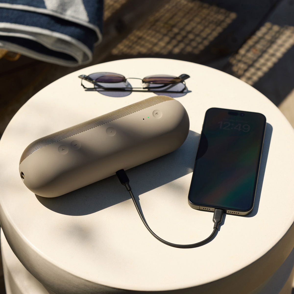 Alt text for this image:
A beige Beats portable speaker sitting on a round table outdoors, charging a smartphone via a connected cable. Sunglasses are also placed on the table, highlighting the speaker’s power-sharing feature in a relaxed outdoor setting.