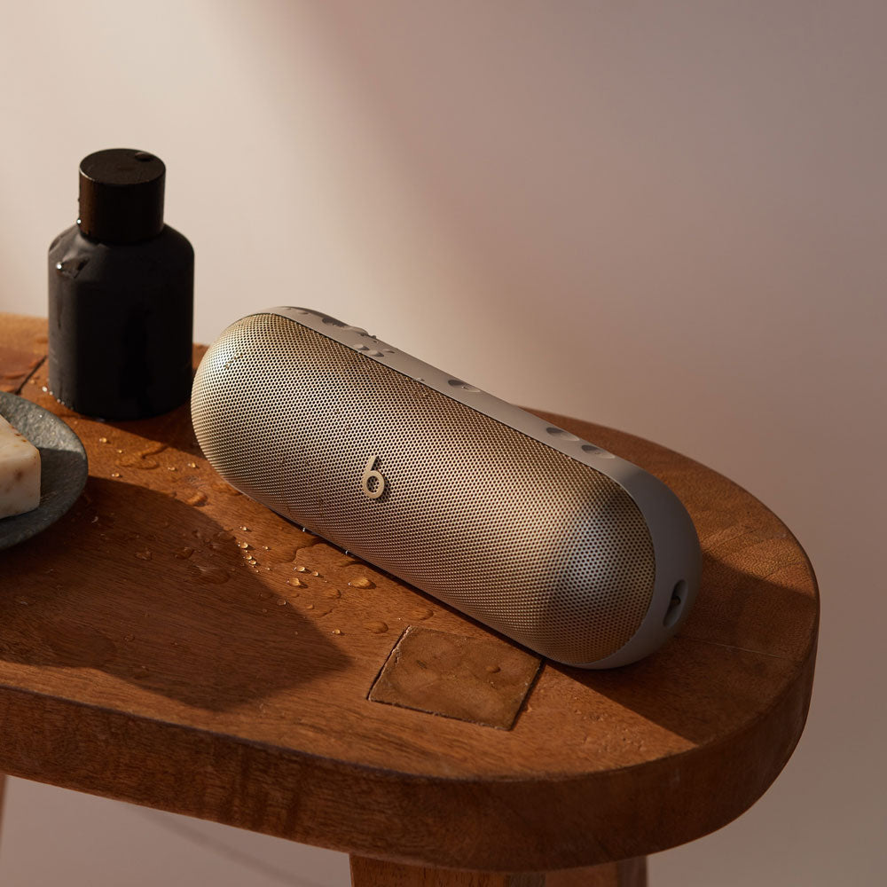 Alt text for this image:
A beige Beats portable speaker sitting on a wooden table, with a few water droplets nearby and a bottle of toiletries, highlighting its water-resistant design and suitability for bathroom or spa settings.