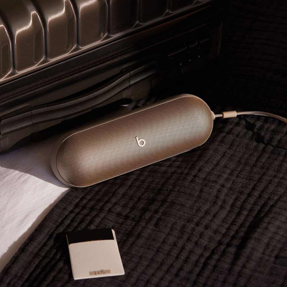 Alt text for this image:
A beige Beats portable speaker placed next to a travel suitcase on a soft black fabric, illustrating its portability and travel-friendly design, with a charging cable connected.