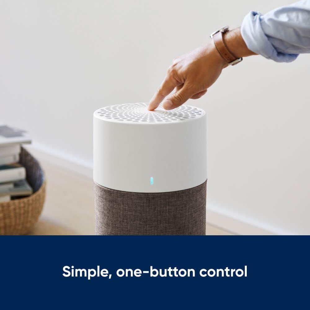 A person presses the top of a modern cylindrical air purifier, demonstrating its simple one-button control. The purifier features a white top with a vented surface and a grey fabric-wrapped lower half. The image includes a caption that reads, "Simple, one-button control."