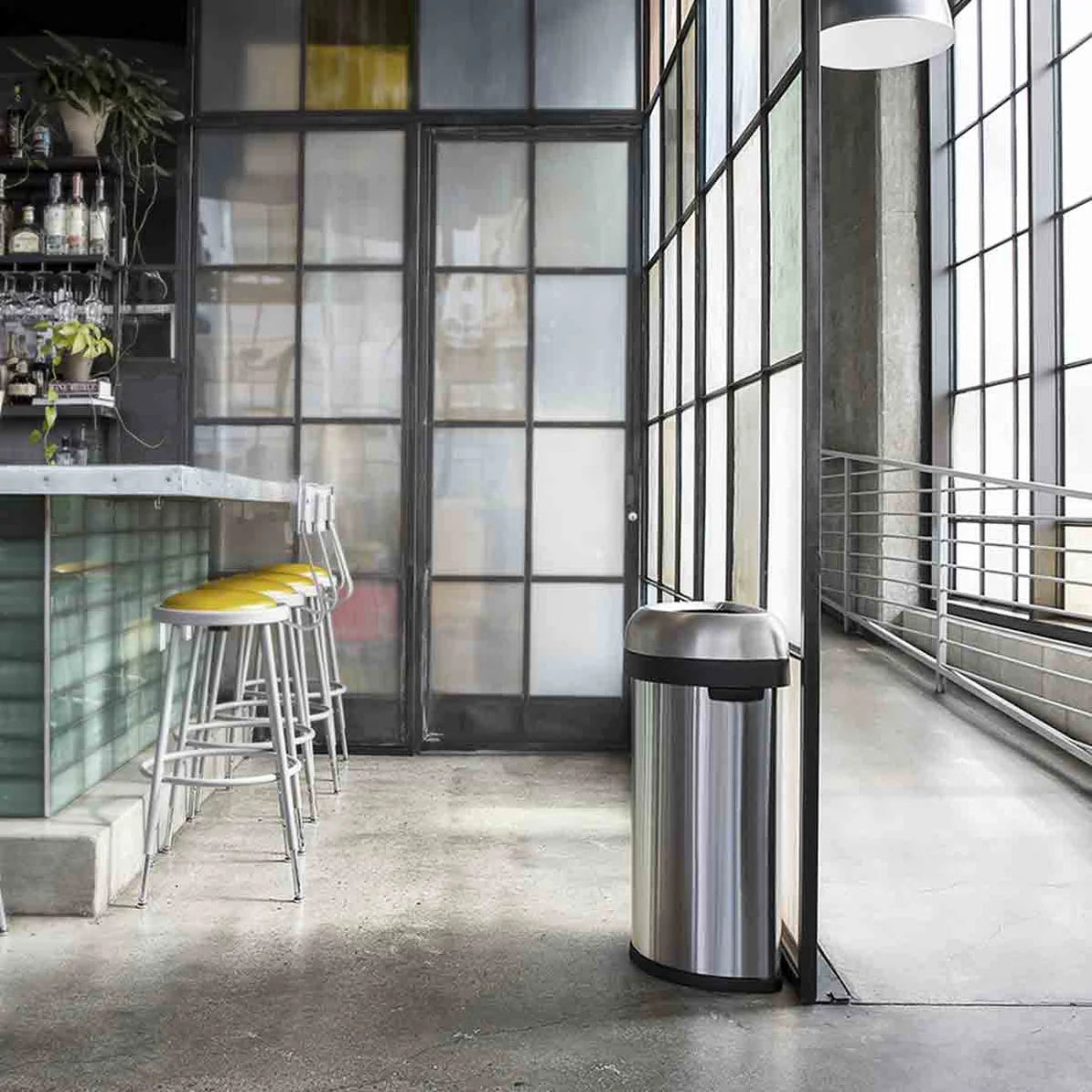 The image shows a modern, industrial-style space with a sleek, stainless steel trash can placed near a bar area. The trash can features a dome-shaped lid with a black trim, blending seamlessly with the contemporary decor. The space has large glass windows, concrete floors, and minimalist furnishings, including bar stools with yellow seats. The trash can's design complements the stylish, clean lines of the environment, making it both a functional and aesthetically pleasing addition to the setting.