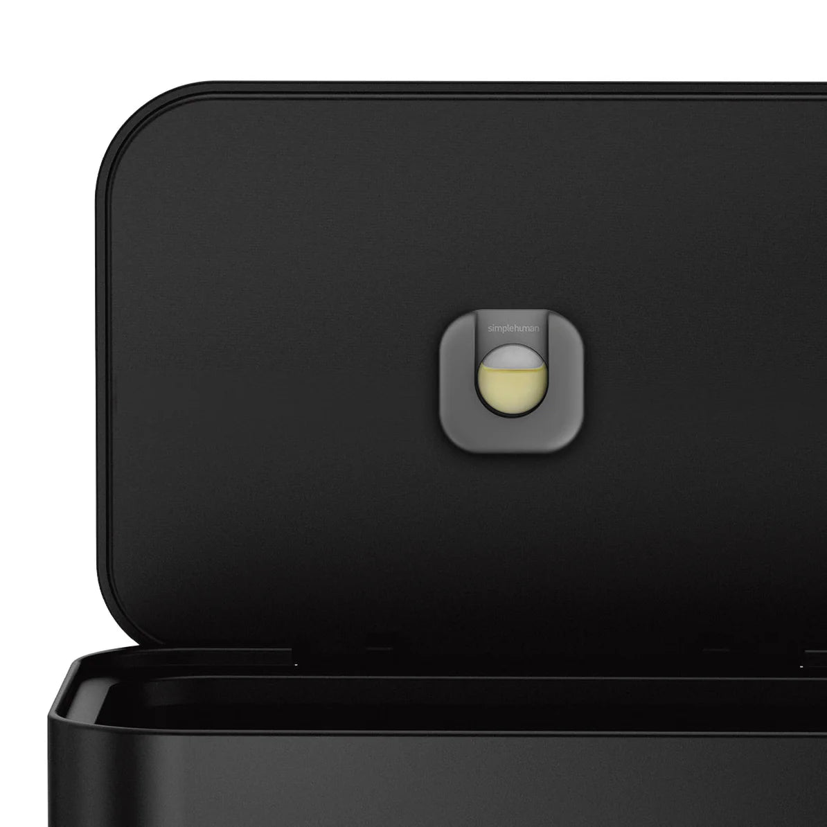The image shows a close-up view of the inside of a Simplehuman trash can lid. The lid is open, revealing a small circular compartment in the center that holds an odor filter. The filter is part of Simplehuman's effort to minimize odors within the trash can, and the compartment is designed to hold a replaceable carbon filter. The Simplehuman logo is subtly visible on the filter holder, and the design is sleek and minimalistic, consistent with the overall modern aesthetic of the trash can. The black interior