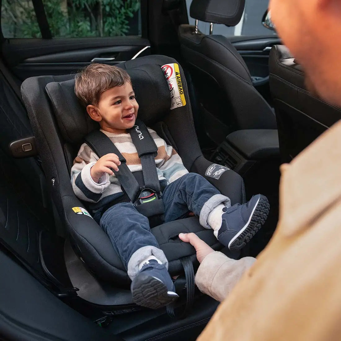 Smiling toddler secured in a rear-facing Nuna convertible car seat with a five-point harness, interacting joyfully with an adult outside the car, highlighting comfort and safety.