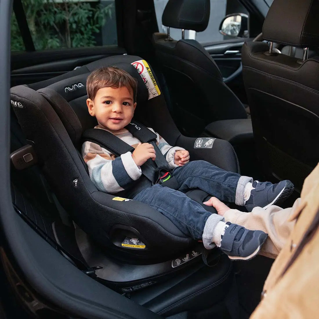 Smiling toddler seated in a rear-facing Nuna convertible car seat inside a car, securely fastened with a five-point harness, while an adult gently holds the child’s legs.