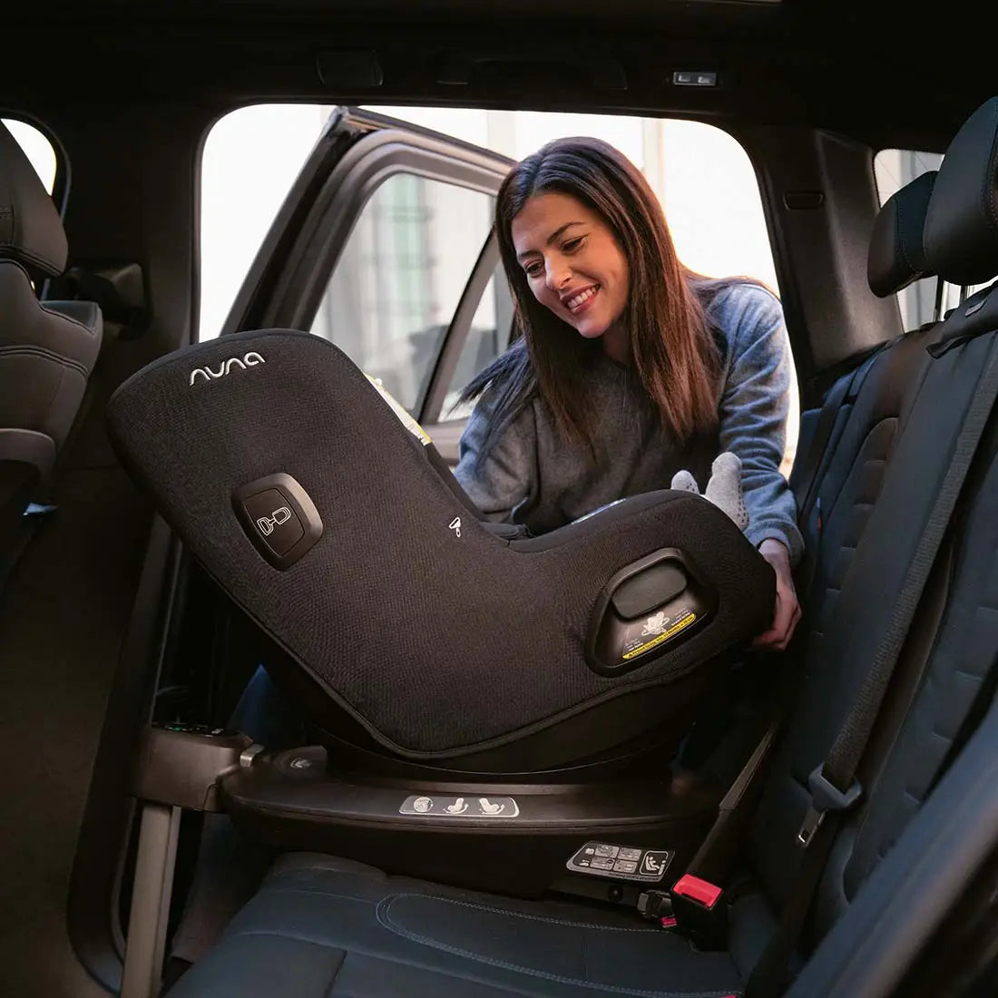 Mother smiling while securing her baby in a Nuna convertible car seat installed in the rear-facing position inside a car, demonstrating safe and comfortable travel setup.