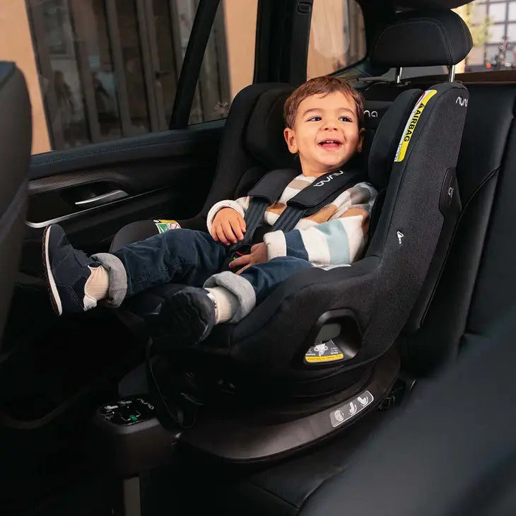 Smiling toddler seated comfortably in a forward-facing Nuna convertible car seat, securely fastened with a five-point harness inside a car, showcasing a safe and joyful travel experience.