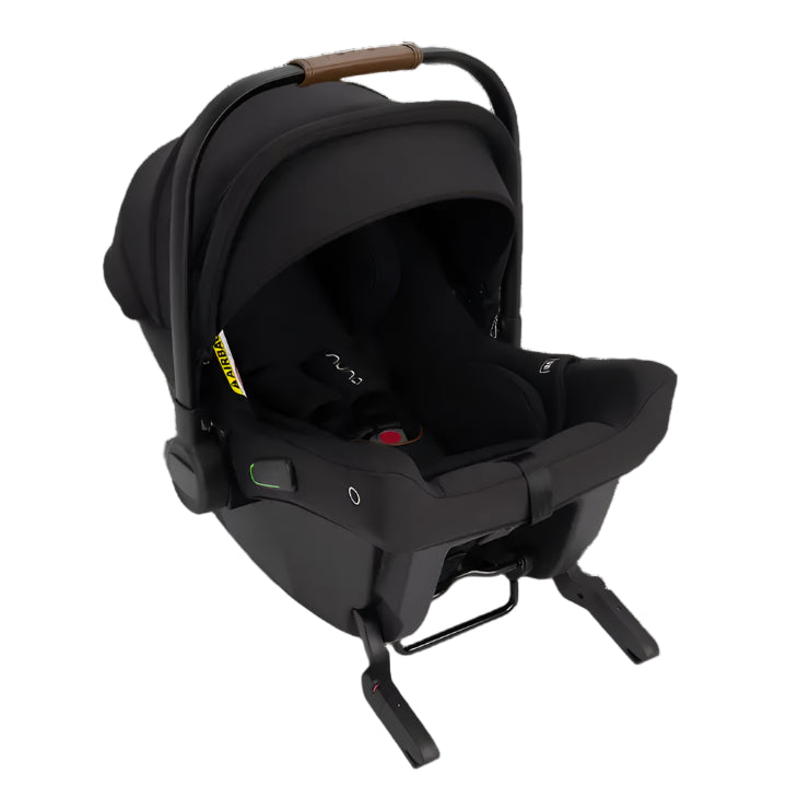 Front three-quarter view of a black Nuna infant car seat with a leatherette handle, extended canopy, and visible ISOFIX connectors. The seat includes a padded five-point harness and a secure base with a load leg for added stability.