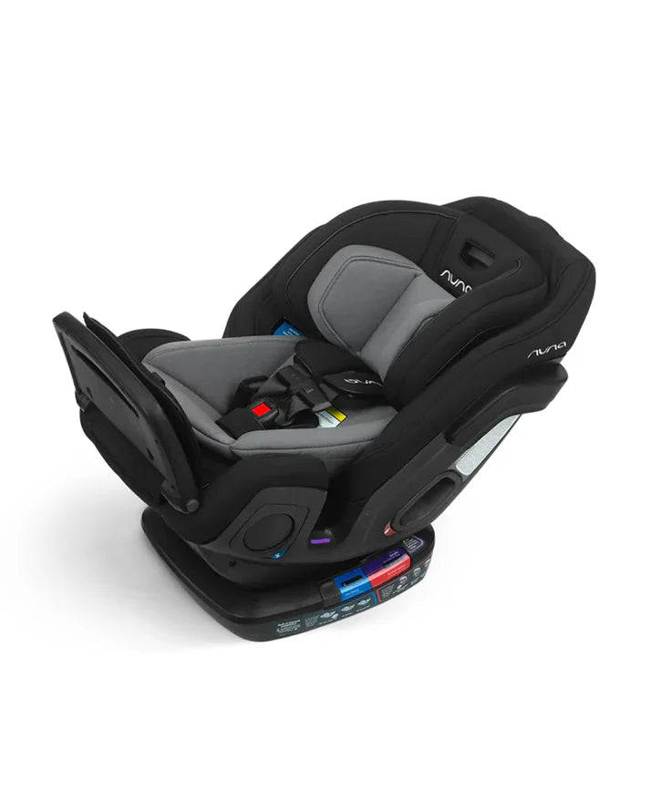 A reclined Nuna convertible car seat in black and grey, showing detailed safety labels, adjustable harness, padded seat, and a leg rest attachment.