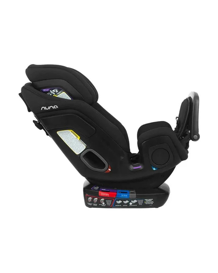 A side view of a black Nuna convertible car seat in a reclined position, showing color-coded installation labels, harness slots, and side impact protection features.