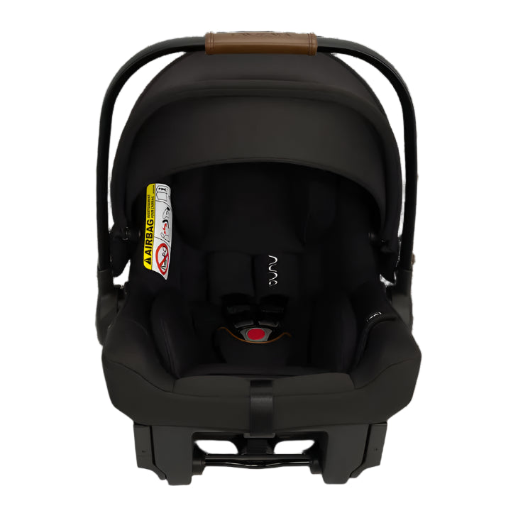 A black Nuna infant car seat with a fully extended canopy, brown leatherette handle, five-point harness, and a visible airbag warning label for rear-facing installation.
