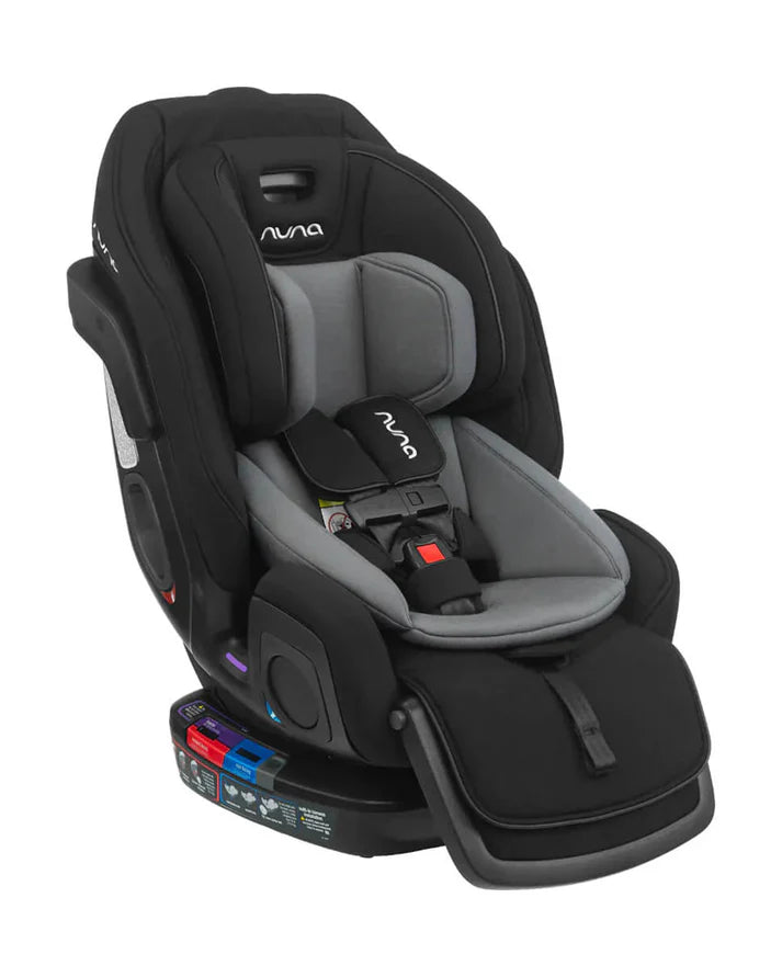 Angled view of a black and grey Nuna convertible car seat featuring a 5-point harness, padded infant inserts, and labeled belt paths for rear- and forward-facing installation.