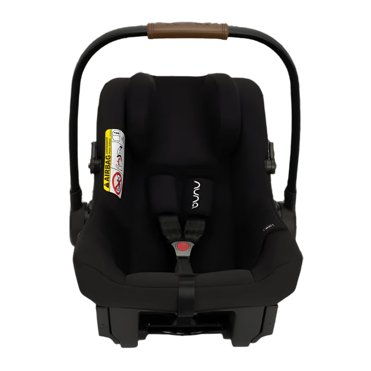 A black Nuna infant car seat with a brown leatherette handle, visible airbag warning label, and secure five-point harness, designed for rear-facing travel.