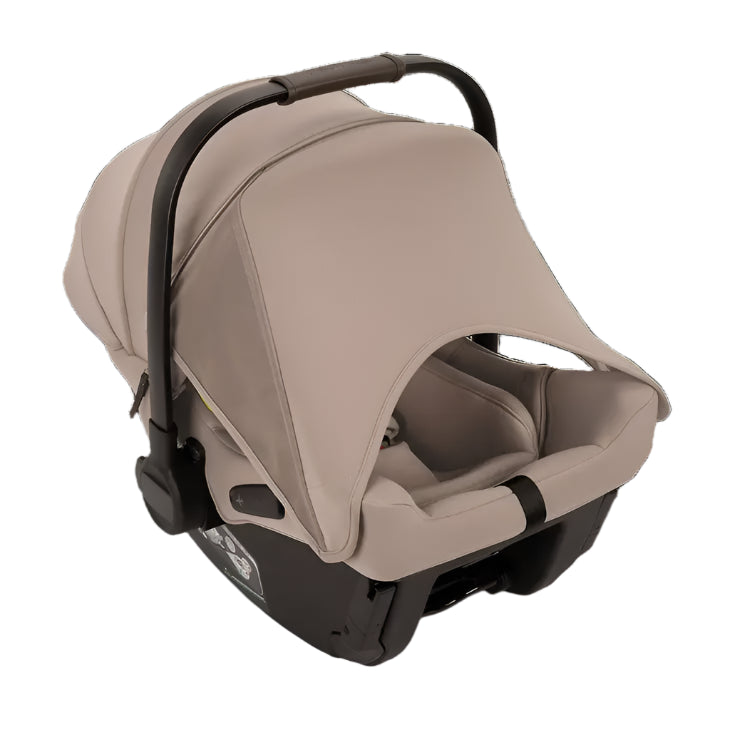 A beige Nuna infant car seat with a fully extended canopy and black carrying handle is shown from a rear side angle, highlighting the ergonomic design and comfortable padded interior.