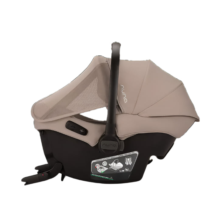 A Nuna infant car seat in beige and black is shown in a side profile with the canopy fully extended, revealing breathable mesh panels and clearly labeled installation instructions on the base.