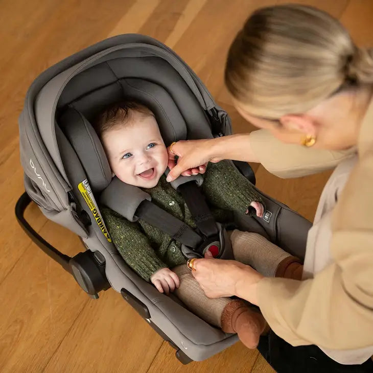 A smiling baby sits securely in a Nuna infant car seat while an adult adjusts the harness, both appearing happy and comfortable on a wooden floor.