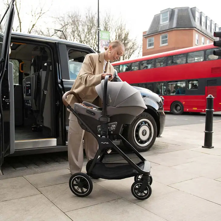 A woman stands beside a black taxi, attaching a Nuna infant car seat to a stroller frame on a city sidewalk, with a red double-decker bus in the background.