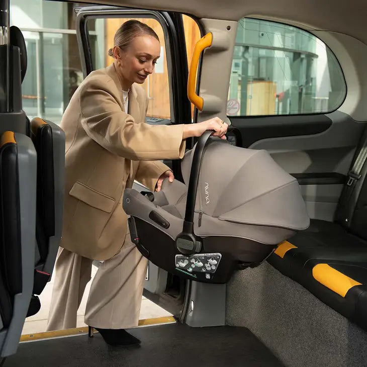 A woman in a beige suit is placing a Nuna infant car seat into the backseat of a taxi, demonstrating its portability and easy installation.