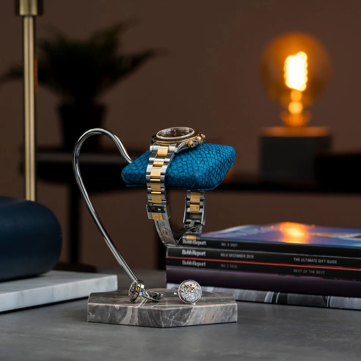 A luxurious wristwatch with a two-tone metal band (gold and silver) and a black dial with gold accents is elegantly displayed on a blue textured cushion. The cushion is mounted on a sleek, curved metal stand with a hexagonal gray and brown veined marble base. The setup includes a pair of cufflinks placed on the base. In the background, there is a stack of books and a softly lit lamp, creating a sophisticated and stylish ambiance.