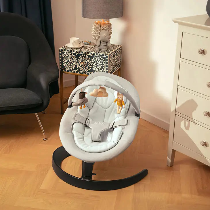 A soft grey baby rocker with plush cushioning and an overhead toy bar featuring a cloud, bunny, and bird toy, placed stylishly in a cozy nursery corner with warm lighting and elegant furniture.