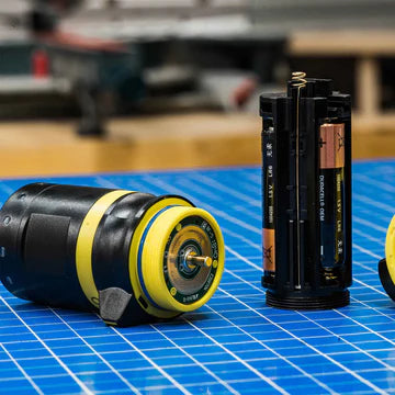 A close-up view of a disassembled flashlight on a blue grid-patterned surface. The flashlight components include the main body, battery holder with AA batteries inserted, and the tail cap. The components are shown separately, revealing the internal structure and design. The main body has a black and yellow color scheme.