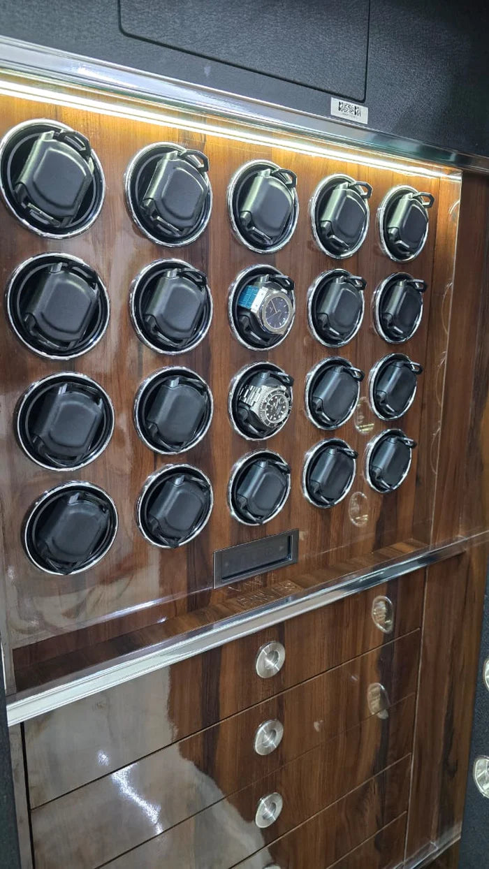 Interior view of a luxurious safe with a wood-grain finish. The top section is equipped with eighteen watch winders arranged in three rows, each capable of holding a single watch. Some winders contain watches, while others are empty. Below the watch winders, there are two drawers with metallic handles, designed for additional storage. The drawers and watch winders are well-lit, highlighting the sophisticated design of the safe's interior.
