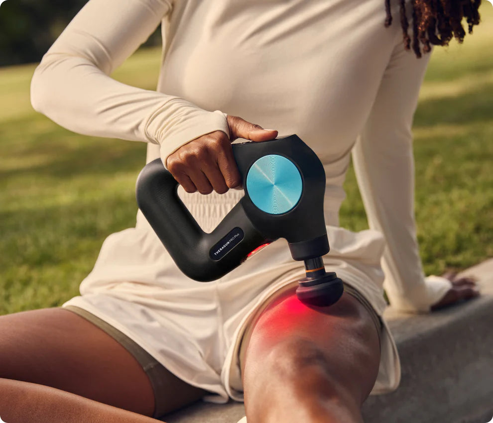A person in a beige long-sleeve top and shorts uses a black and blue massage gun on their thigh while sitting outdoors. The massage gun emits a red light on the targeted muscle area.